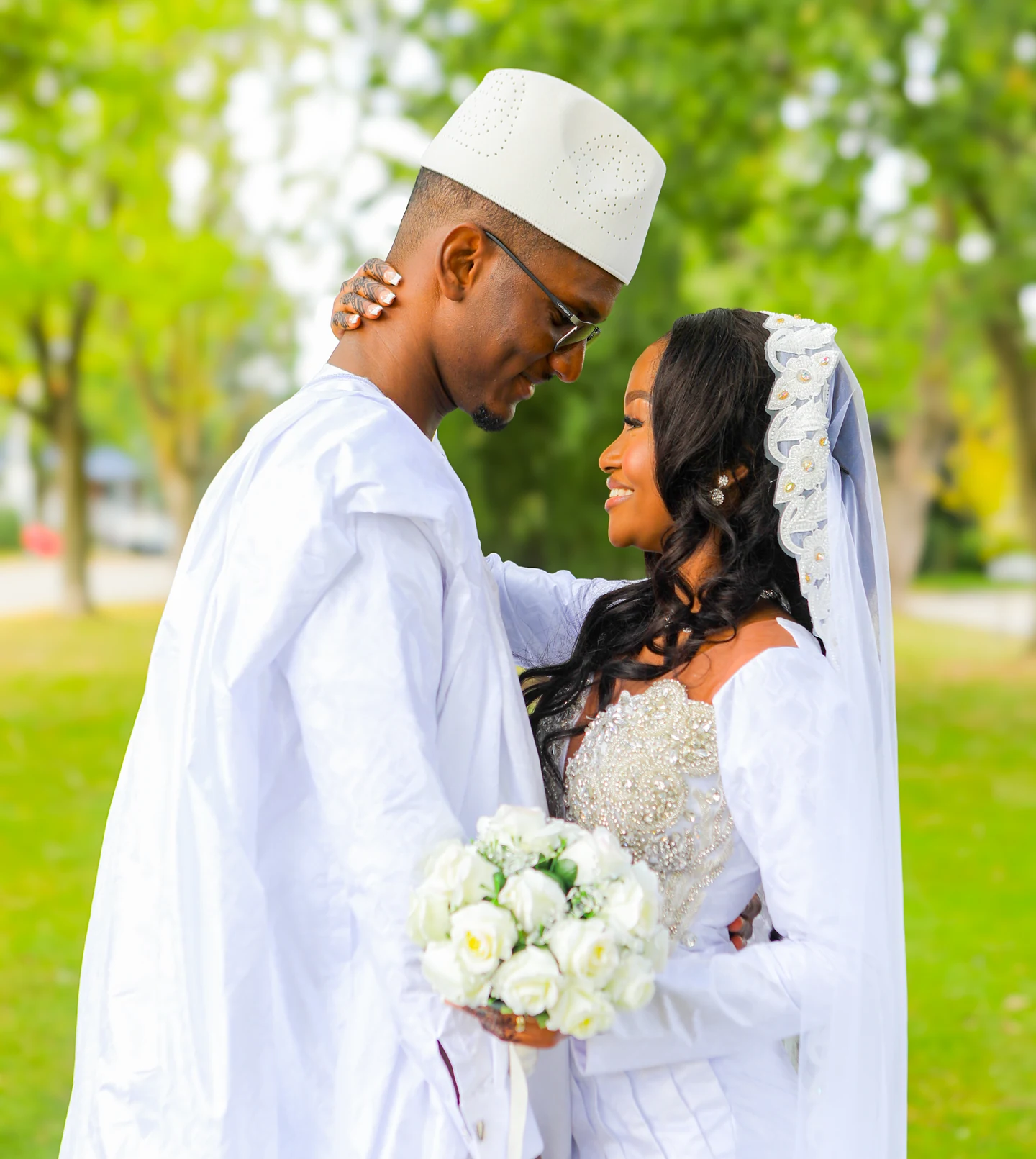 Un couple de mariés en tenues blanches échange un regard souriant, avec un bouquet, dans un parc.